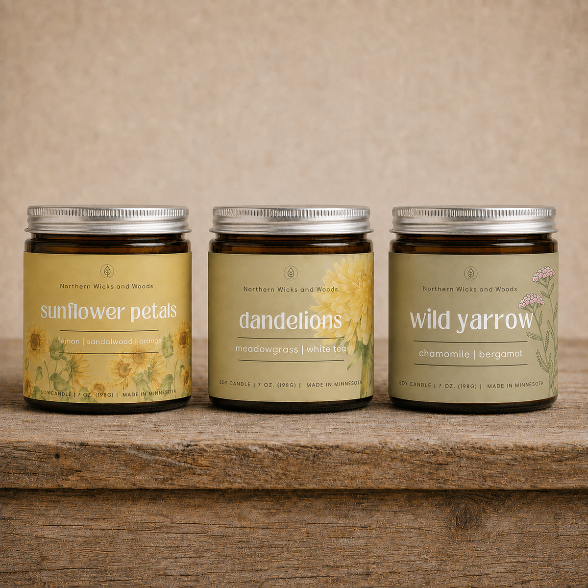 Three amber glass candle jars with silver lids from Northern Wicks and Woods, labeled 'sunflower petals,' 'dandelions,' and 'wild yarrow,' arranged on a rustic wooden surface against a beige textured backdrop.