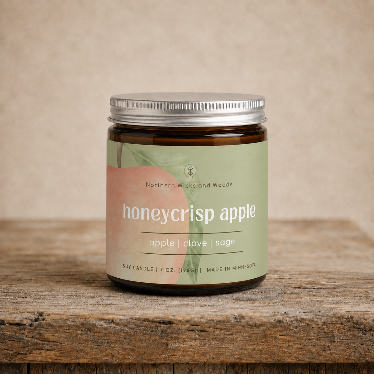A 7oz amber glass jar candle with a silver metal lid and a light green label reading 'honeycrisp apple' by Northern Wicks and Woods, sitting on a rustic wooden surface against a neutral beige background.