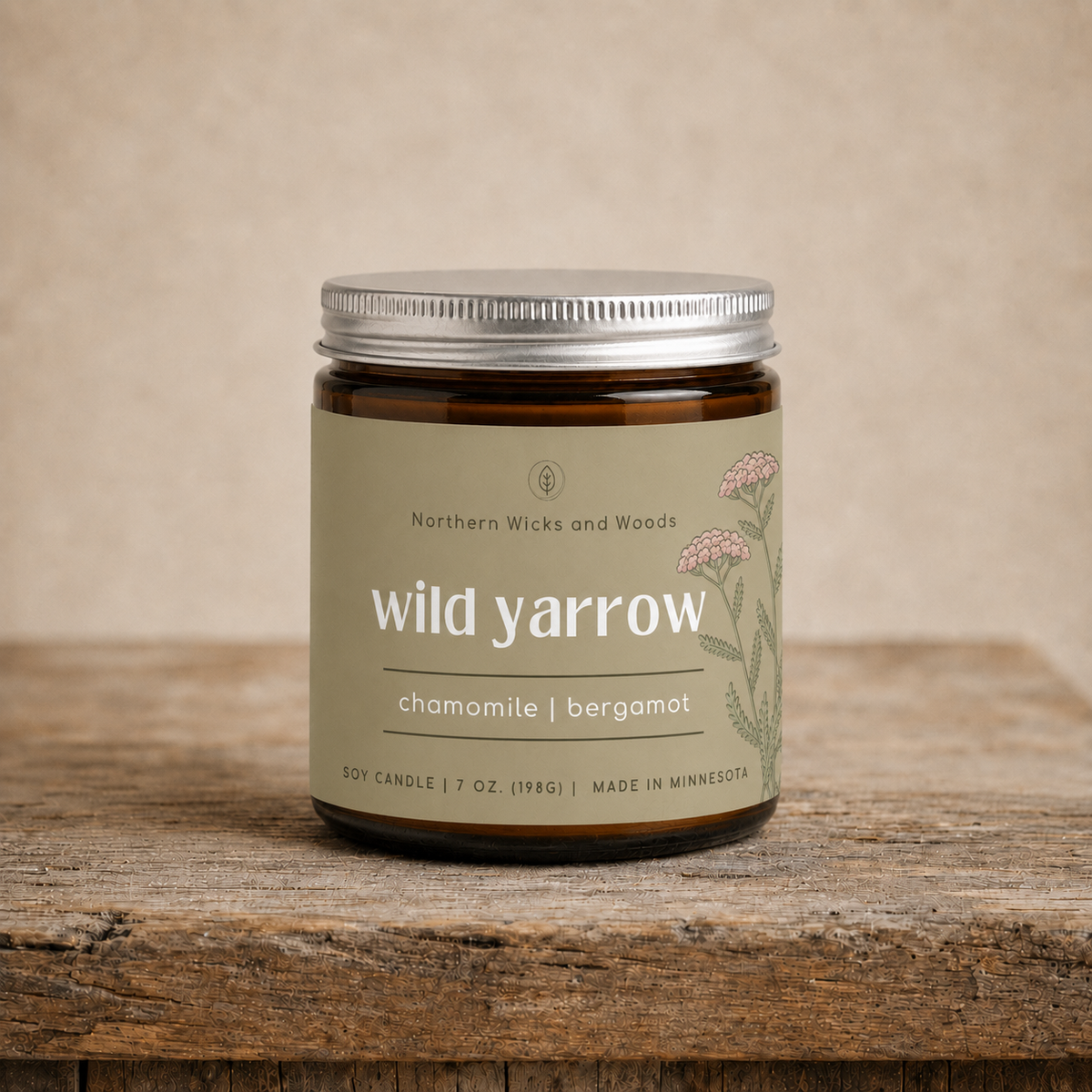 A soy candle in an amber glass jar with a silver metal lid and a beige label reading 'wild yarrow' and 'chamomile | bergamot,' set on a rustic wooden surface against a neutral beige background.