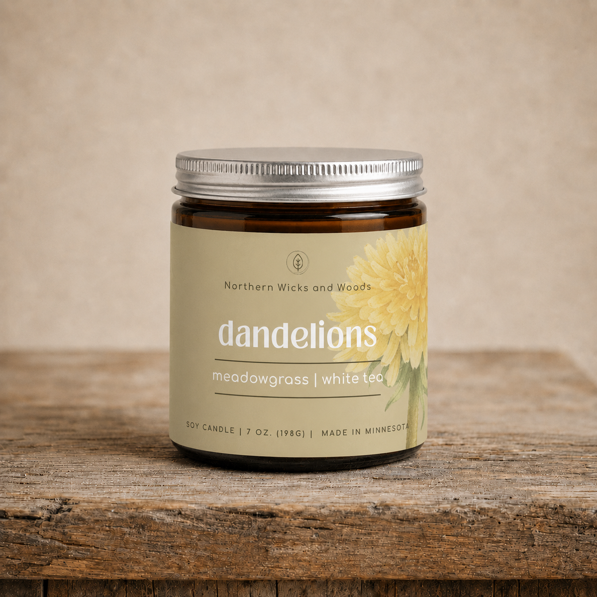 A soy candle in a dark amber glass jar with a silver metal lid, featuring a beige label with a dandelion illustration and the brand name 'Northern Wicks and Woods'. The candle is displayed on a rustic wooden surface against a neutral beige background.