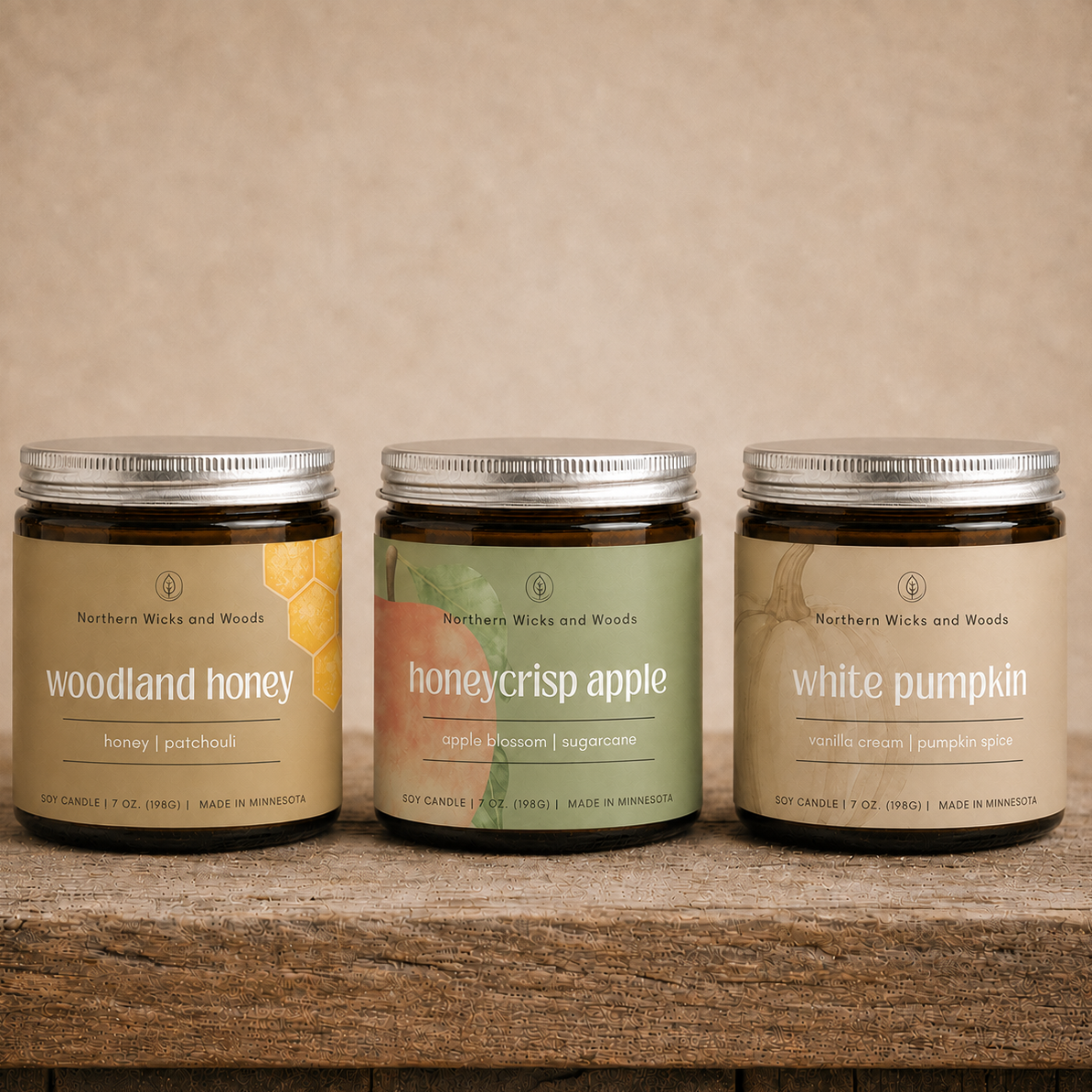 Three glass jars of Northern Wicks and Woods beeswax candles are arranged on a rustic wooden surface against a textured beige background. The jars are labeled 'woodland honey', 'honeycrisp apple', and 'white pumpkin'.